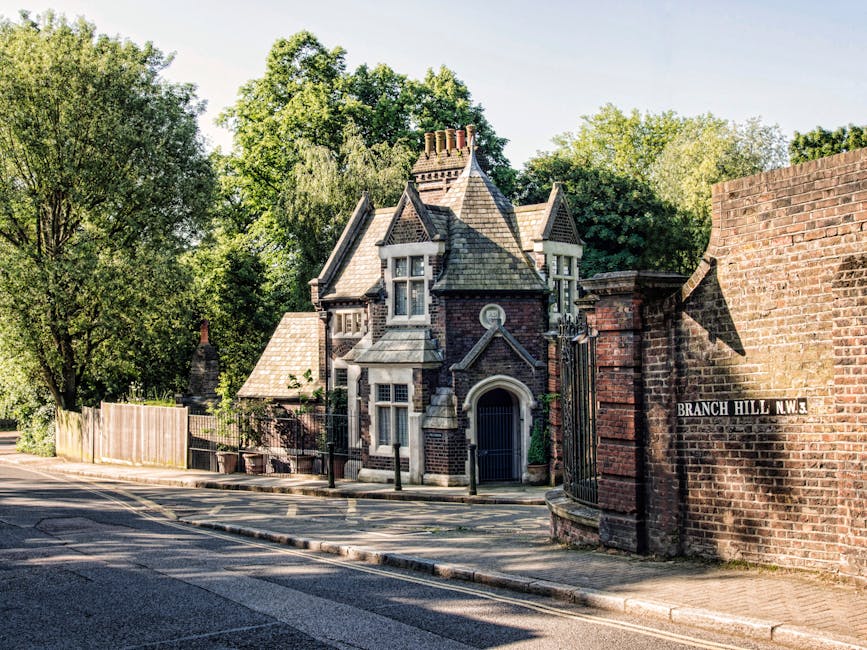 A narrow residential street with a historic, Victorian-style house featuring a steeply pitched roof, ornate chimneys, and decorative stonework situated behind a brick wall and black wrought iron gate. The house has tall, narrow windows with white frames and is partially obscured by mature green trees and foliage. To the right, a street sign reads 'Branch Hill N.W.3', indicating the location. The pavement and road are visible in the foreground, with shadows cast by the trees creating a dappled pattern on the ground. This scene may be relevant to house removals, illustrating the type of property involved in local furniture transport and packing and moving logistics handled by companies like Swiss Cottage Removals.