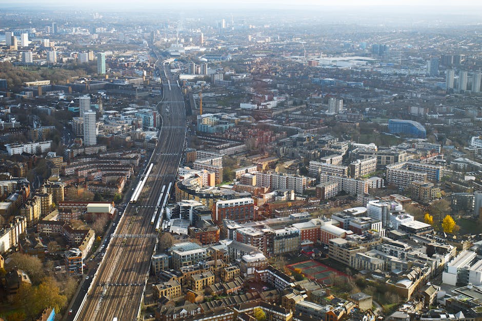 An aerial view of a densely populated urban area showing a railway line running through the cityscape, surrounded by a variety of residential and commercial buildings of different architectural styles and heights. The railway tracks are visible in the center of the image, with trains stationary on the tracks. Nearby, there are apartment blocks, office buildings, and some green spaces with trees. The environment appears to be captured during daylight hours with clear visibility, illustrating the scale of the city. This scene reflects typical urban logistics and transport infrastructure, relevant to house removals and furniture transport services provided by Swiss Cottage Removals, especially when coordinating moving and packing processes near homes and apartments in Swiss Cottage near Finchley Road station.